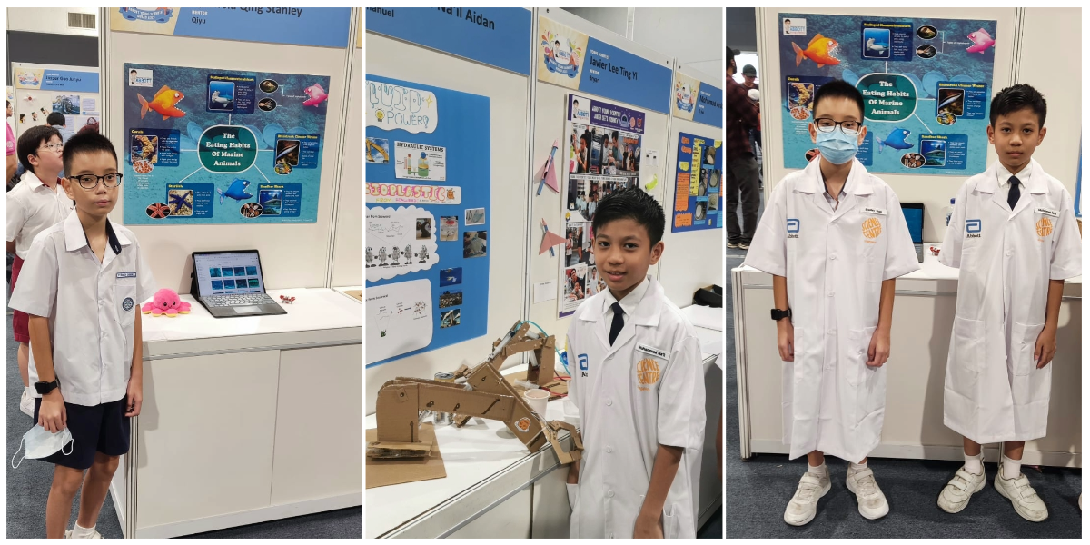 Young students in white lab coats stand before colorful science fair project boards about marine animals and hydraulic systems.